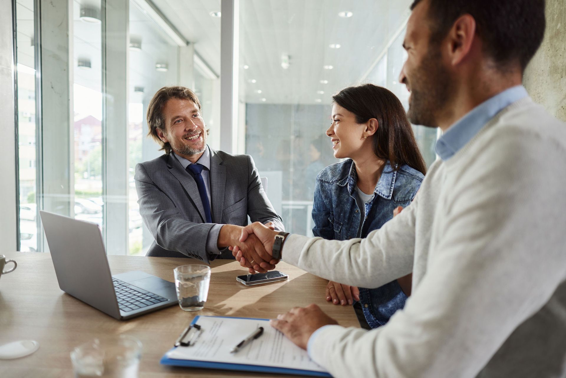 Two men shake hands over a table with a woman smiling. Laptop and documents present in a modern office. Two men shake hands over a table with a woman smiling. Laptop and documents present in a modern office.