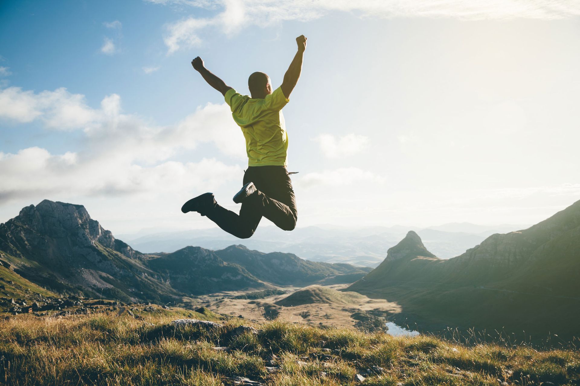 Man jumps for joy on mountain top, arms raised, sunny sky, scenic landscape.