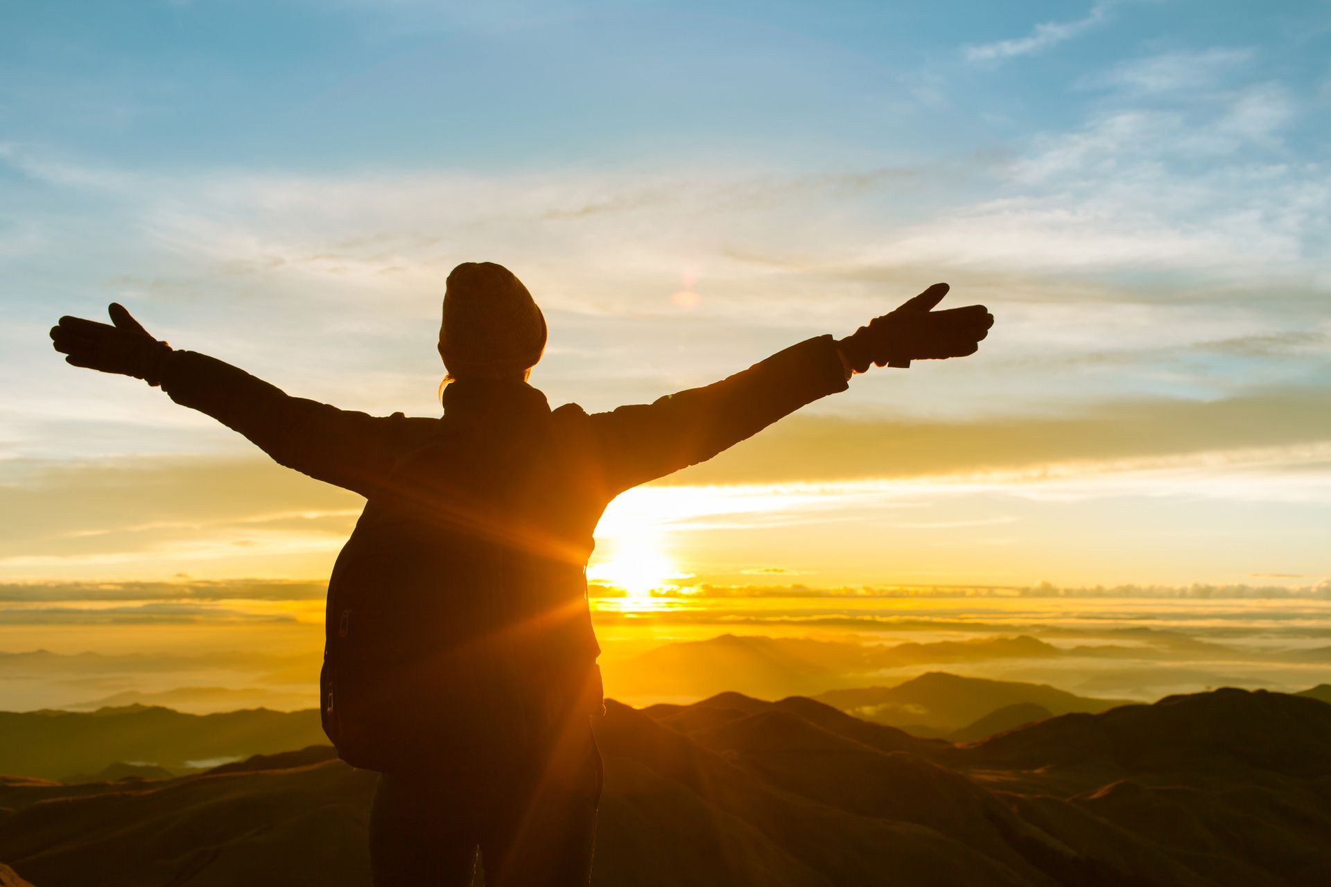 Silhouette of person with arms outstretched on mountain, sun rising.