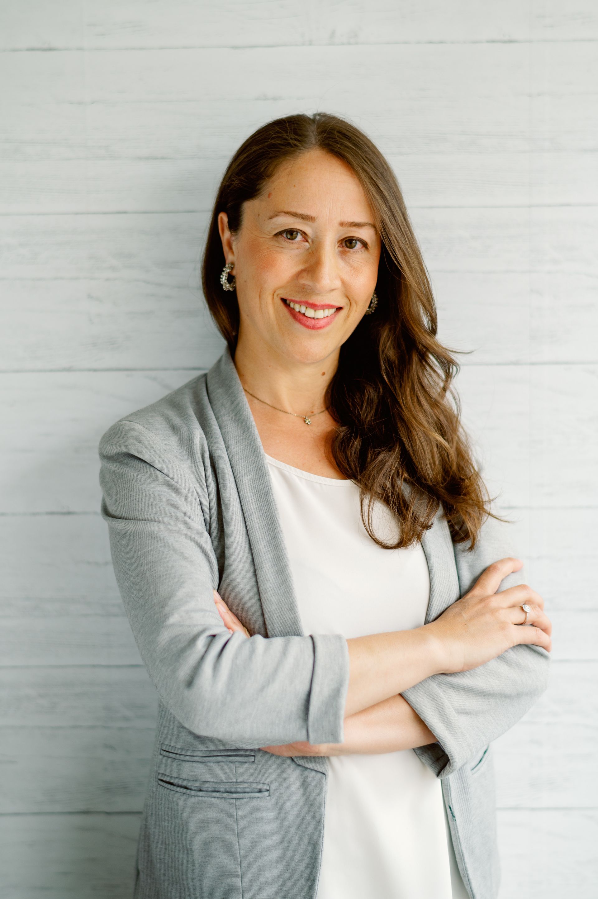 A woman in a grey jacket and white dress is standing with her arms crossed and smiling.