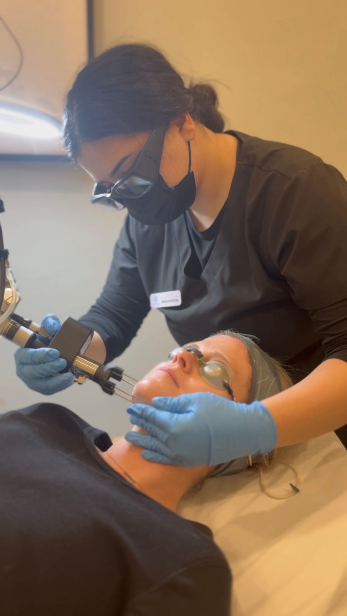 A woman is wearing a mask and gloves while a man is getting a laser treatment on his face.