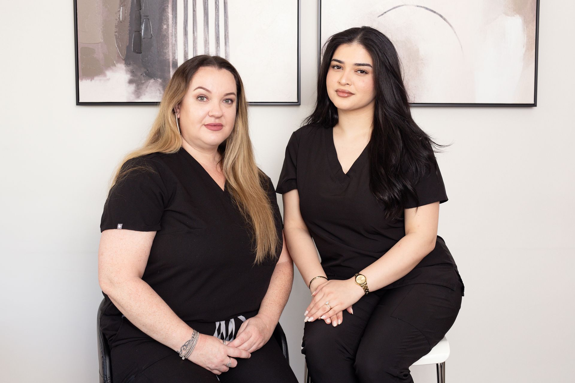 Two women in black scrubs are sitting next to each other in front of a wall.