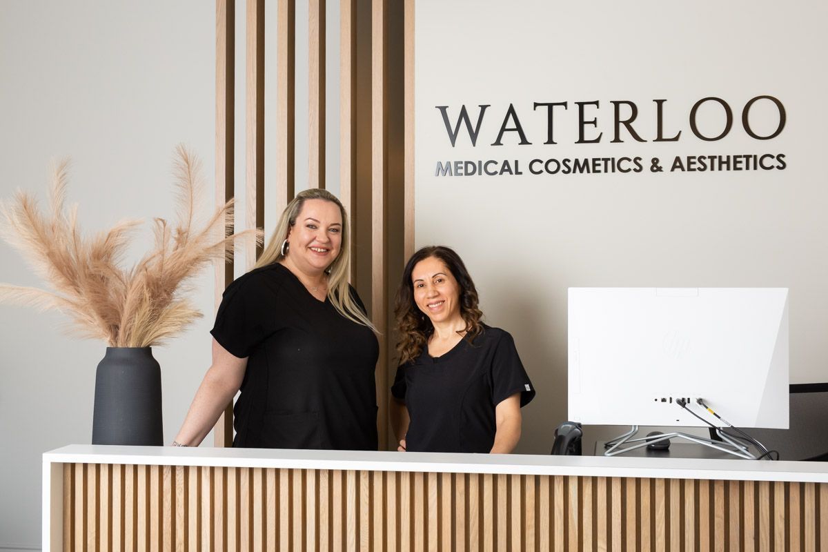 Two women are standing at a desk in front of a sign that says waterloo medical cosmetics and aesthetics.