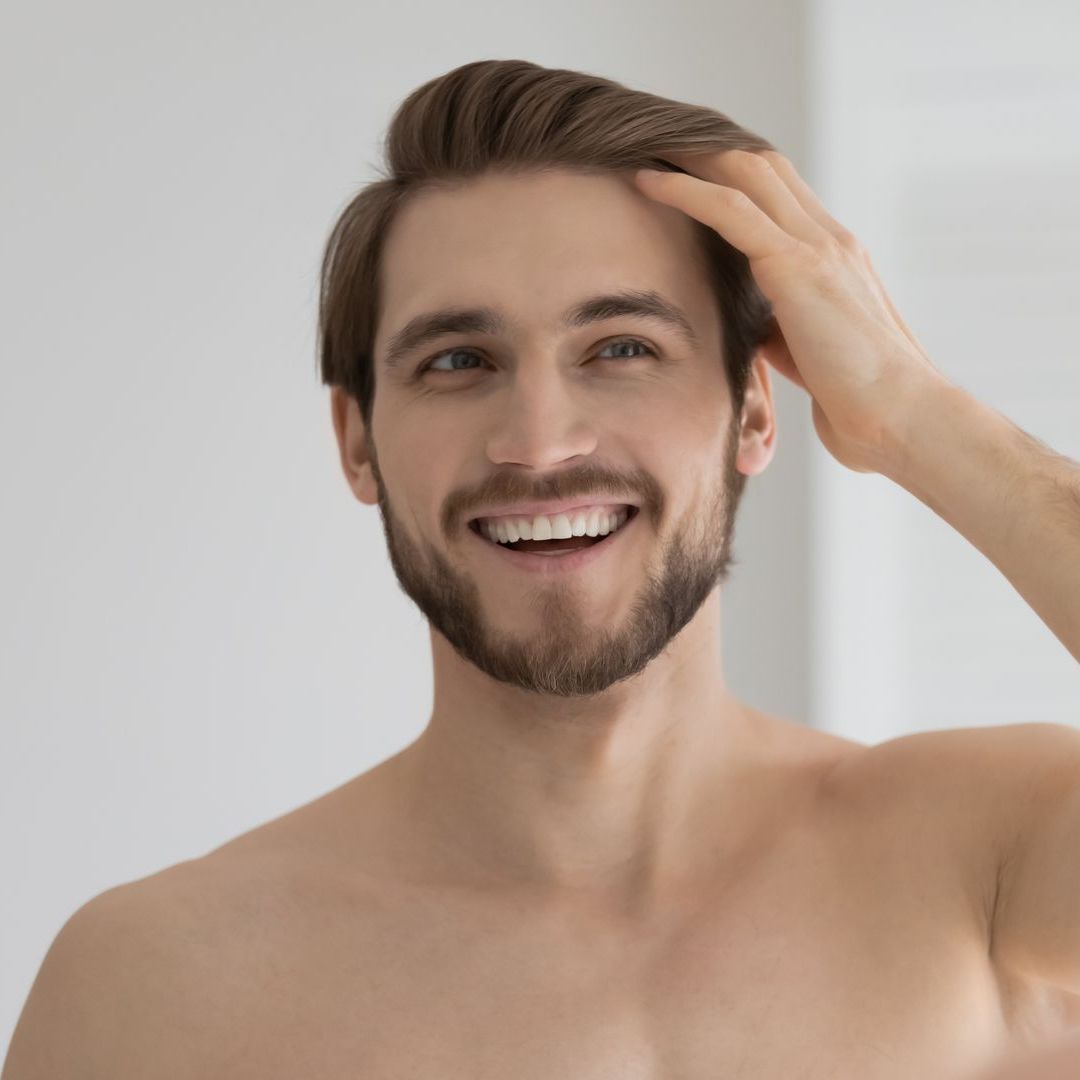 A shirtless man is looking at his hair in the mirror.