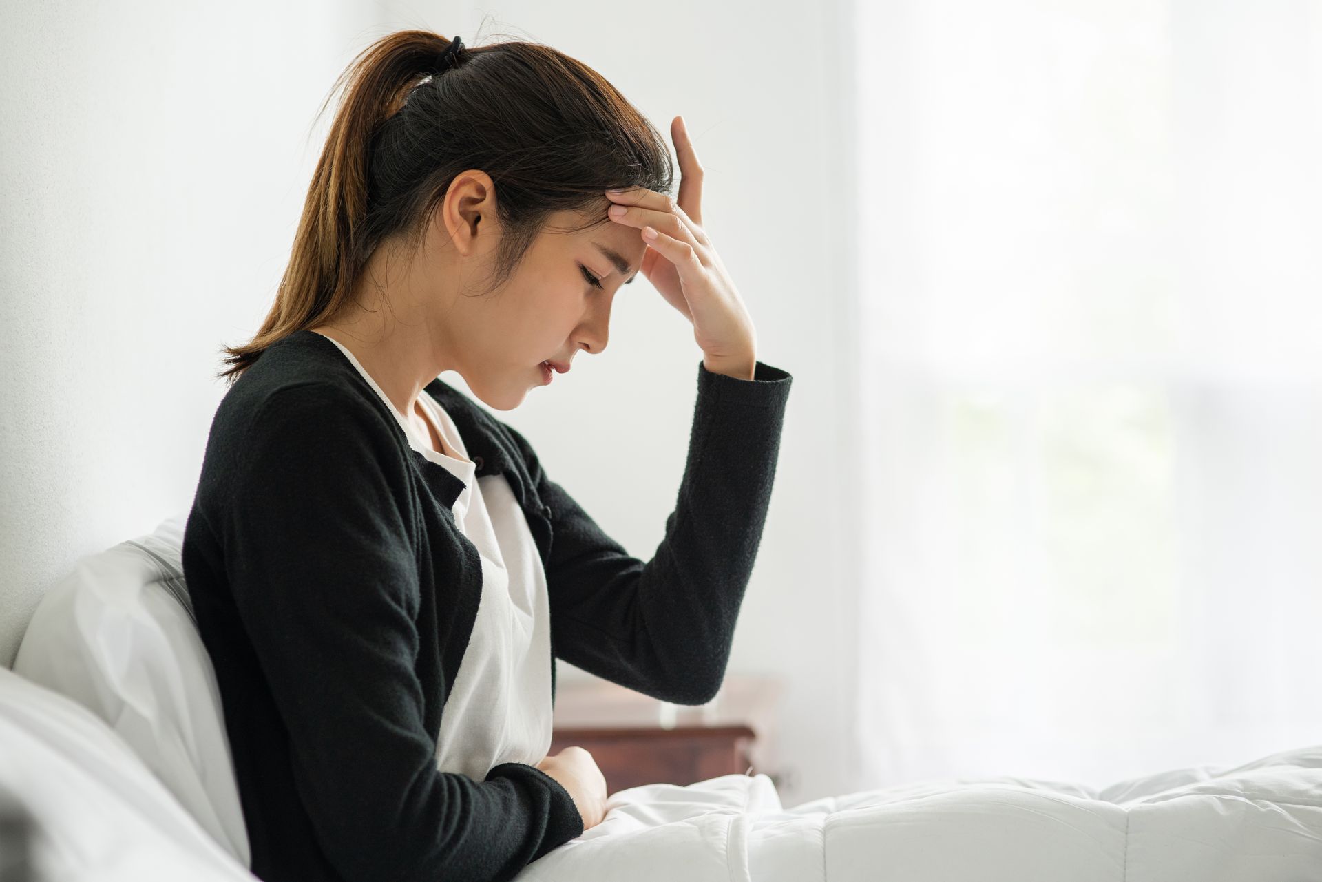 A woman is sitting on a bed with her hand on her forehead.