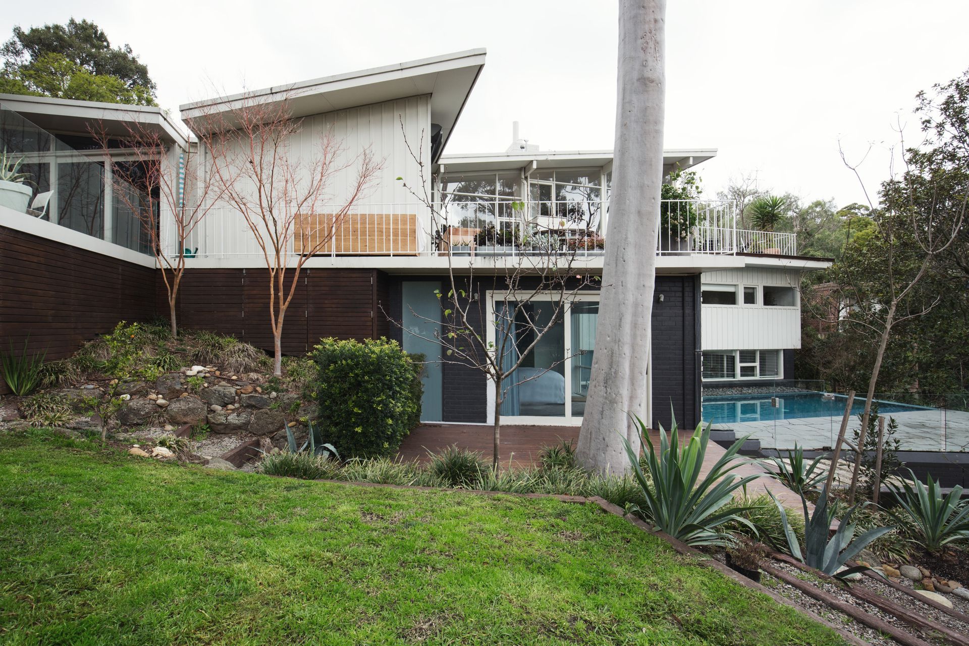 Modern multi-level home with pool on a hillside, featuring brick and white exterior, large windows, and a sloping roof.