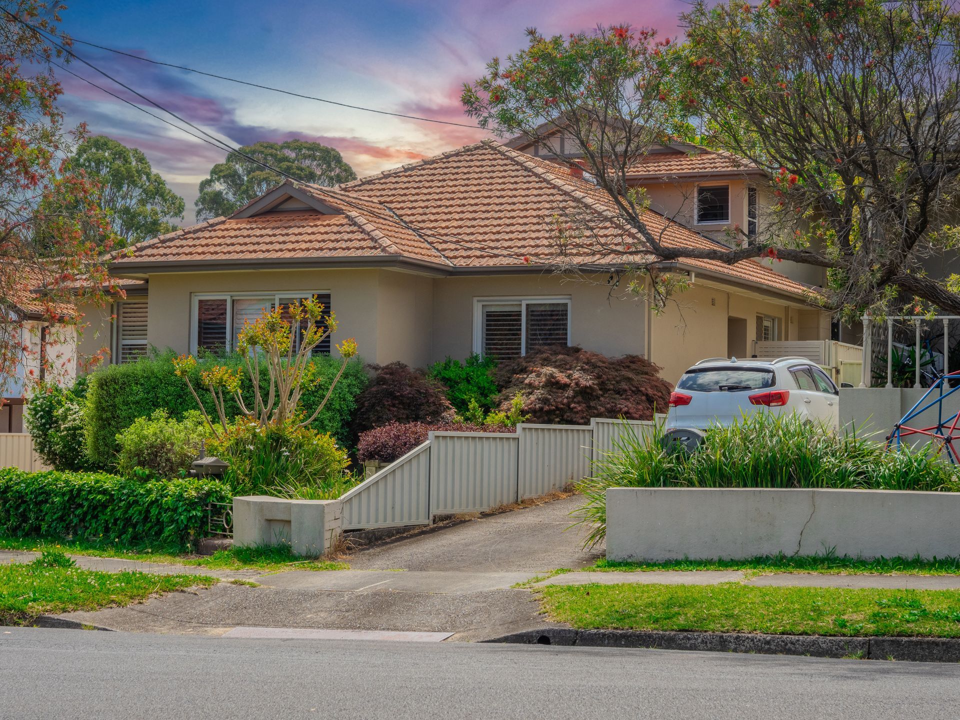 Suburban house with tan stucco, red-tiled roof, white car in driveway, green lawn, and vibrant sunset sky.