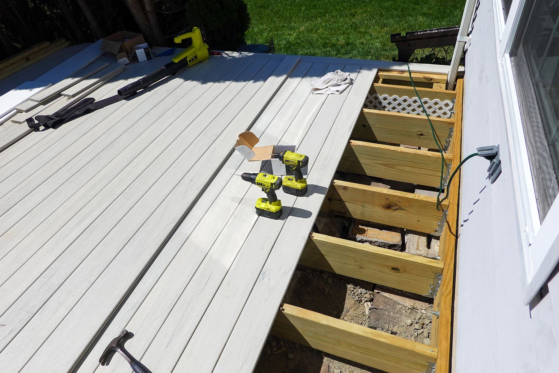 Deck construction; gray composite boards laid, wooden frame, power tools, near house with window.