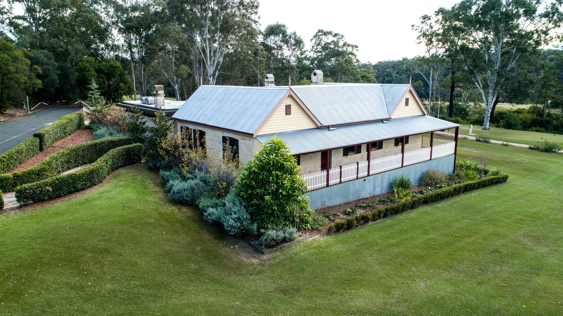 A long, light-colored house with a tin roof sits in a grassy yard, surrounded by gardens and trees.