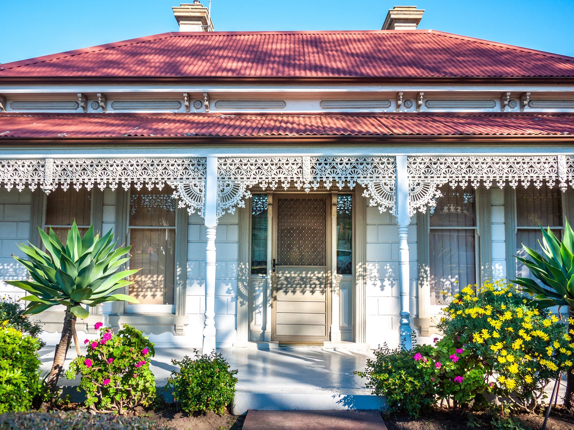 White house with ornate porch trim, red roof, and lush landscaping against a clear blue sky.