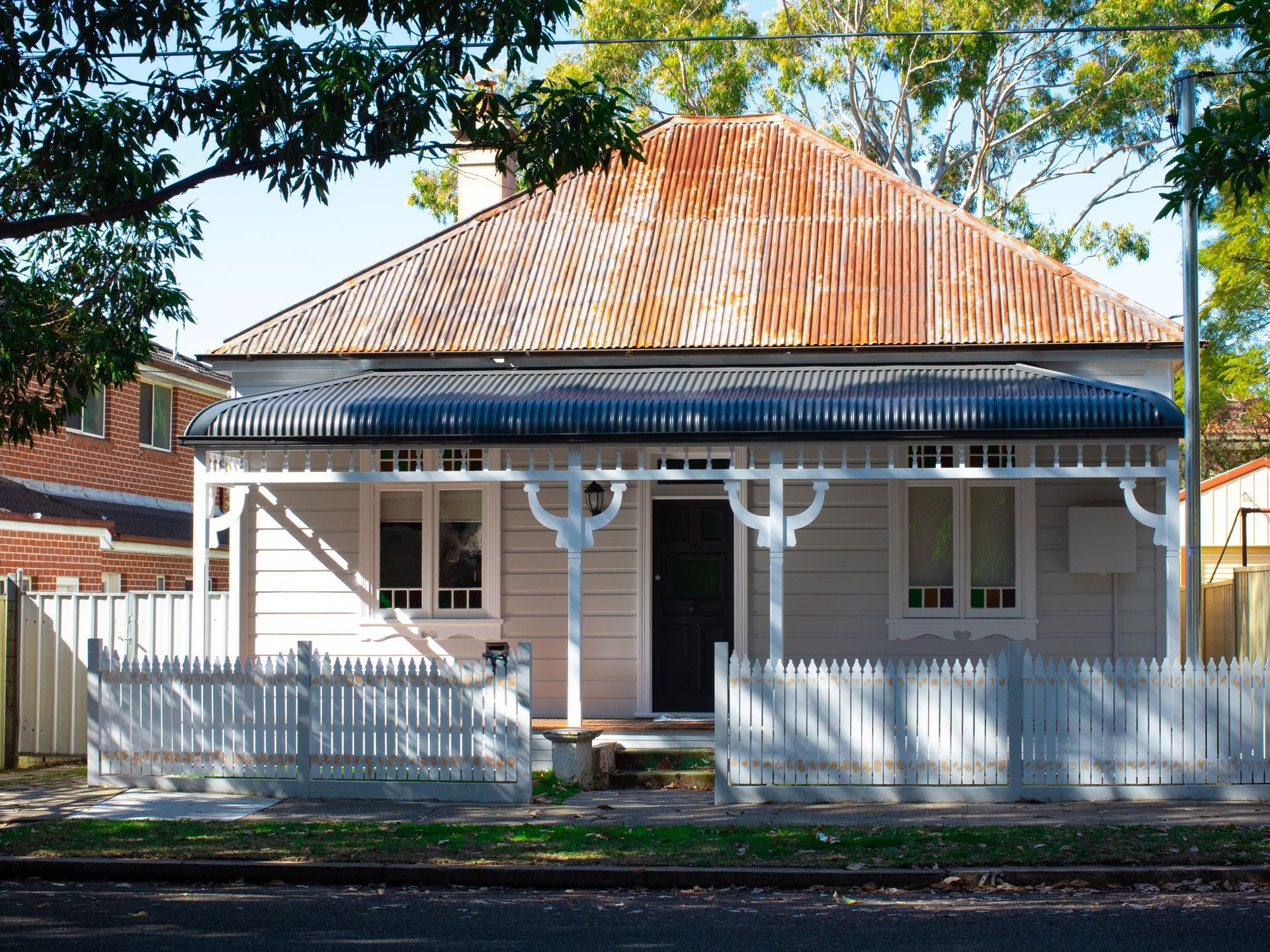 White cottage with rusty roof, porch, and picket fence, trees in the background.