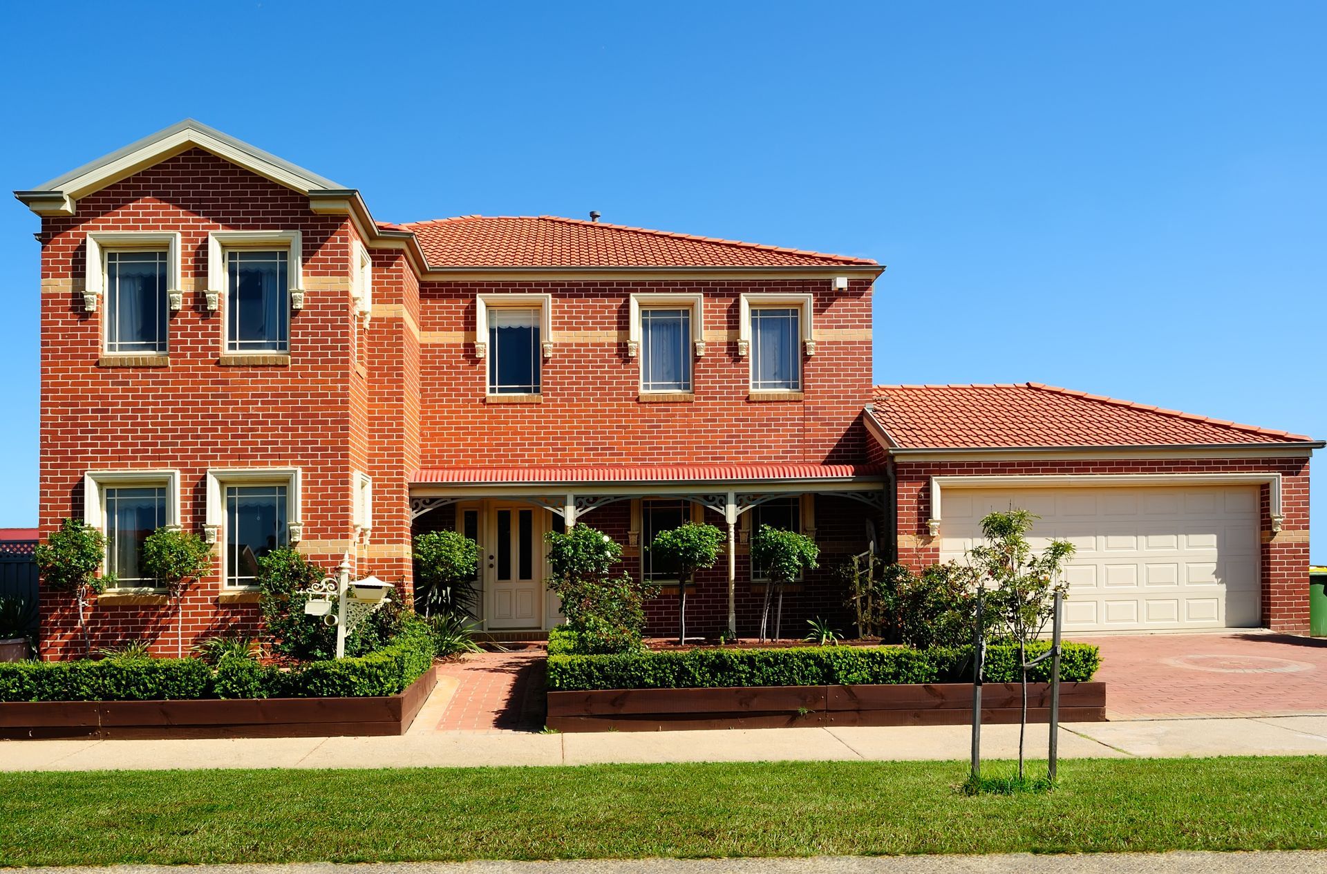 Two-story brick house with a red tile roof, white garage door, and manicured landscaping under a blue sky.