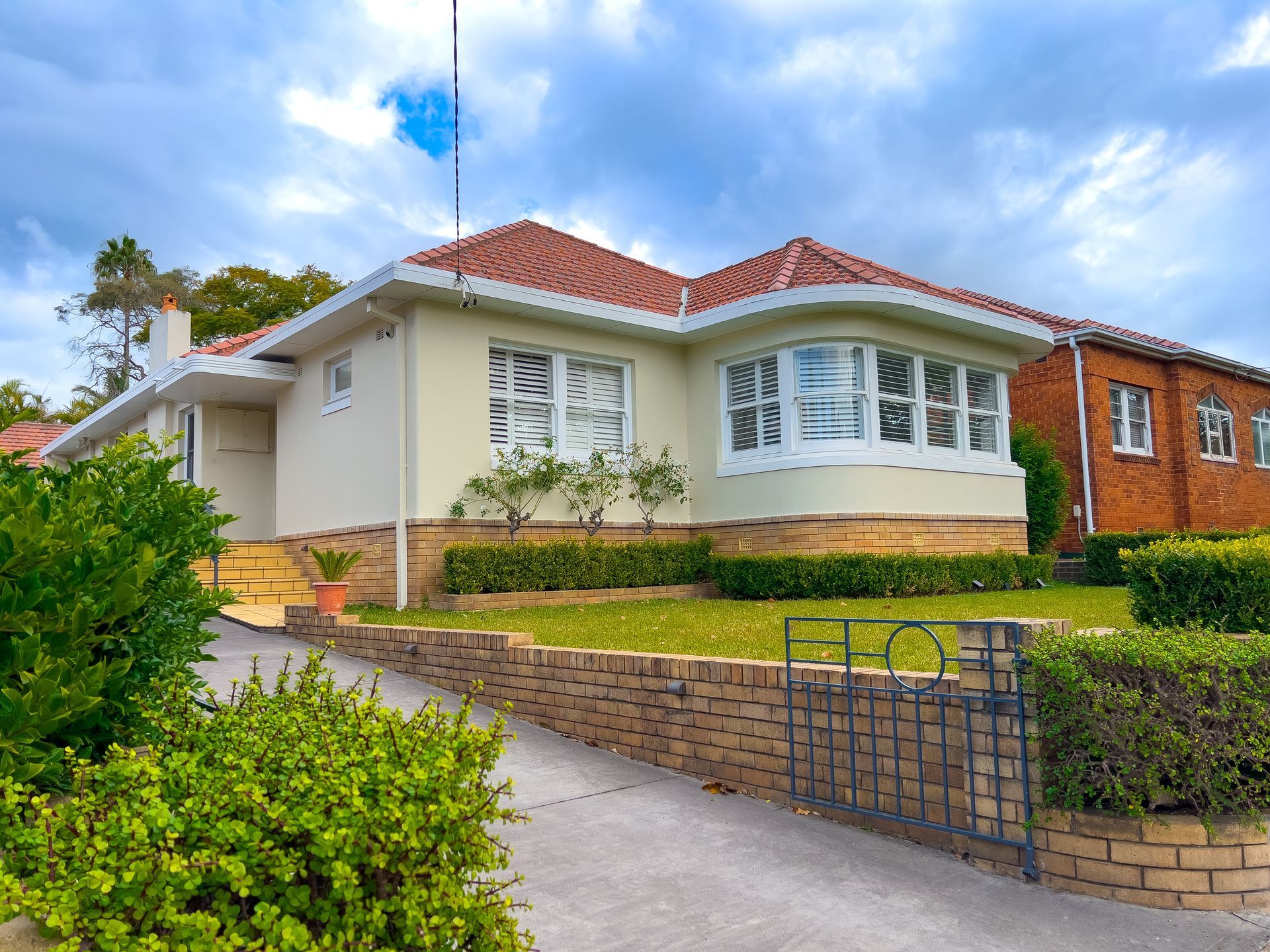 Beige house with a red-tiled roof, bay window, and manicured lawn, fronted by a brick wall and driveway.