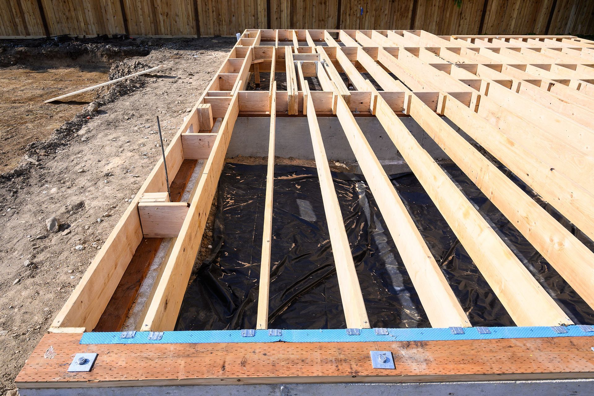 Wooden floor framing under construction, outdoors. Lumber on a concrete base, plastic sheeting visible.