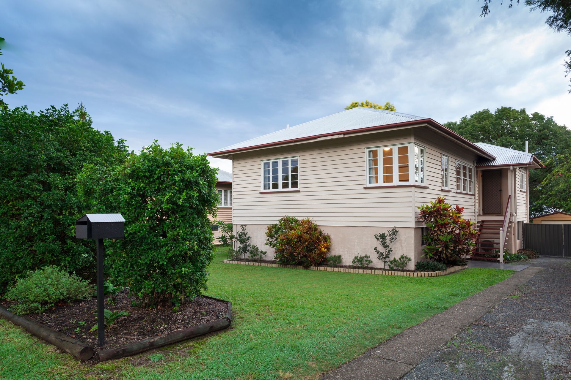 Beige bungalow house with a green lawn, mailbox, and gravel driveway under a cloudy sky.