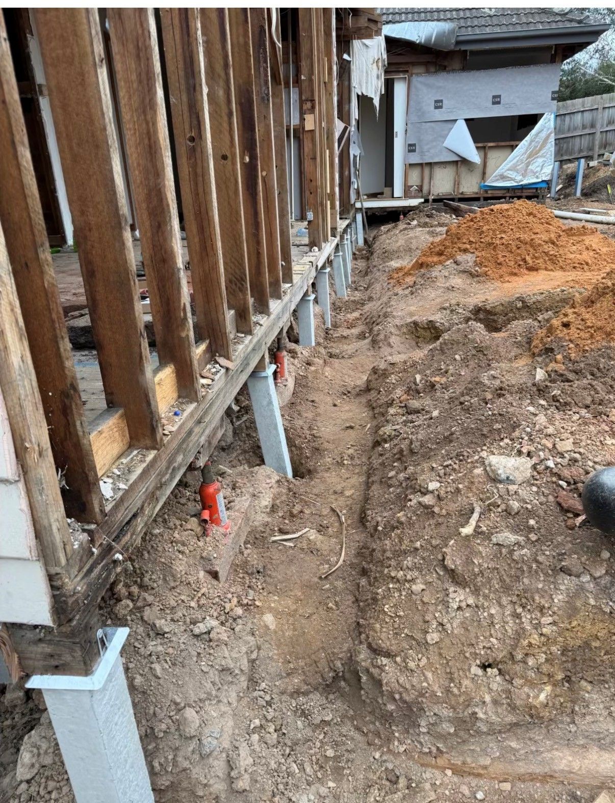 Construction site: wooden structure supported by metal posts in a dirt trench, red pipes visible.