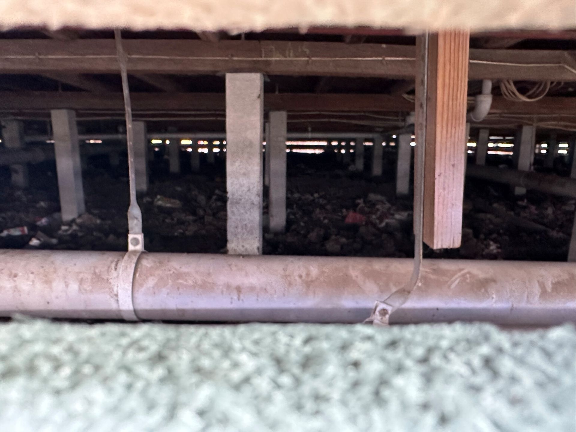 View from beneath a house. Wooden beams, concrete posts, and plumbing are visible among debris and dirt.