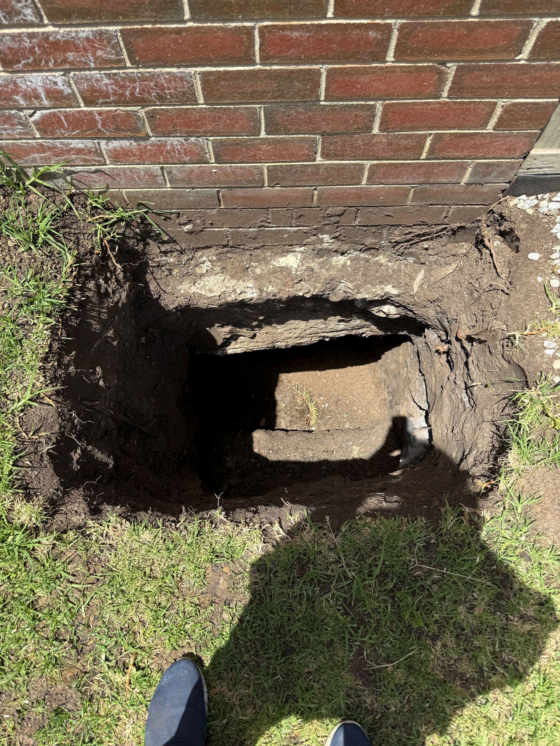 Hole dug in the ground near a brick wall, revealing a concrete block. Grass and dirt surround.