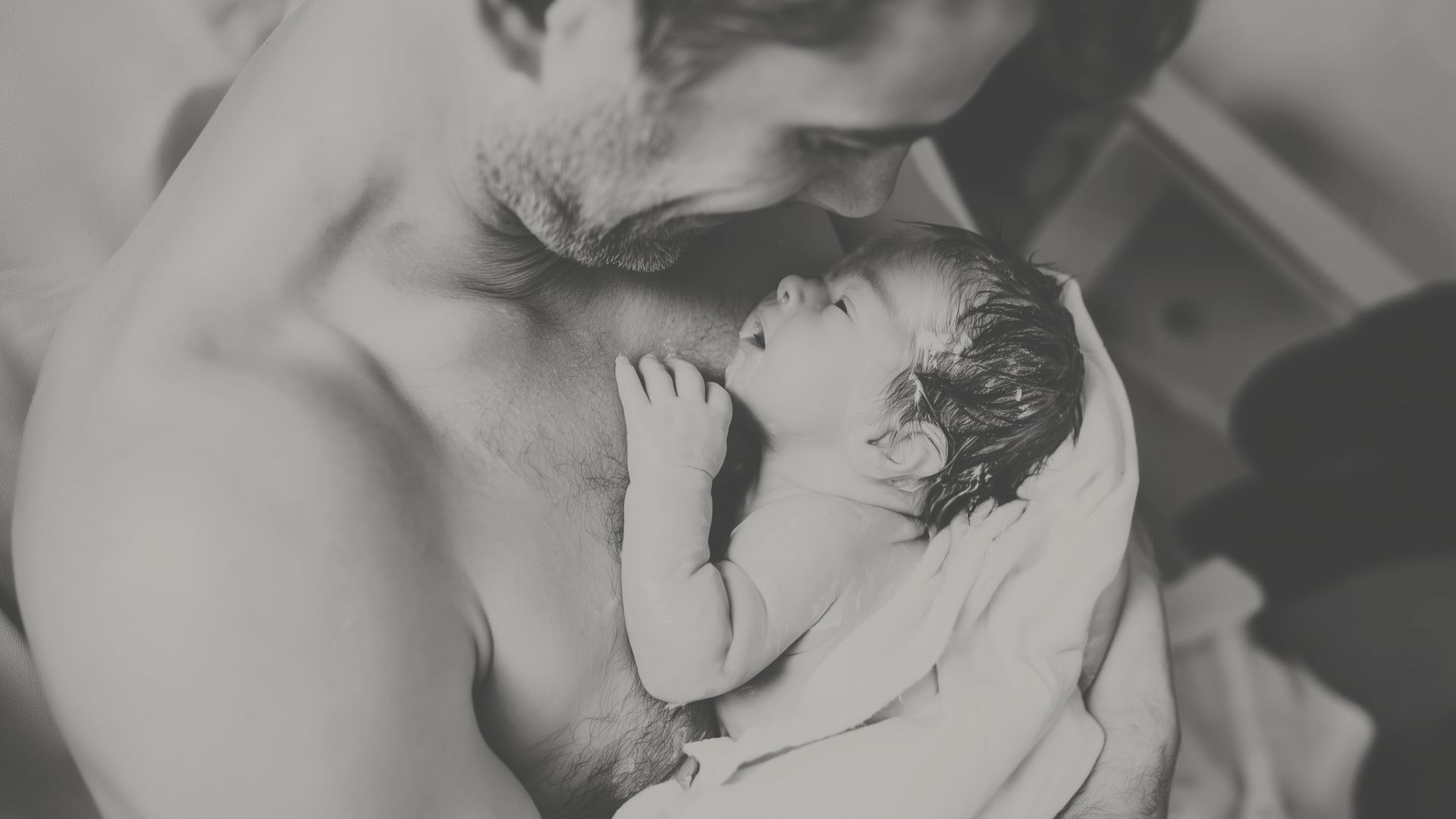 Father gazes lovingly at newborn baby wrapped in a towel against his bare chest. Black and white.