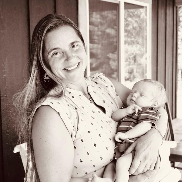 Woman with long hair smiles holding a sleeping baby; outdoors on porch.