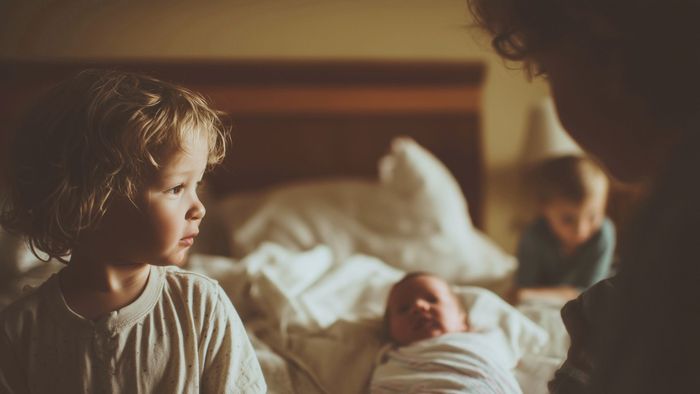 Three children gathered around a newborn on a bed. Soft lighting, blonde hair, focused gazes.