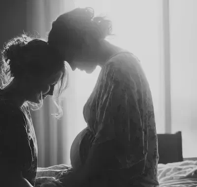 Pregnant woman and friend, in soft light, share a tender moment.