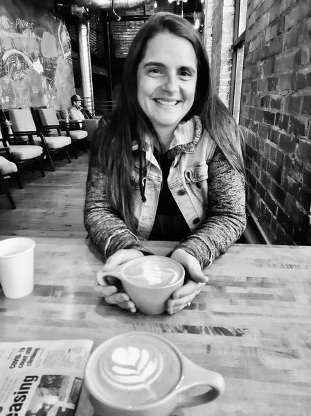 Woman smiling, holding coffee cup with latte art. Seated at table, in cafe.