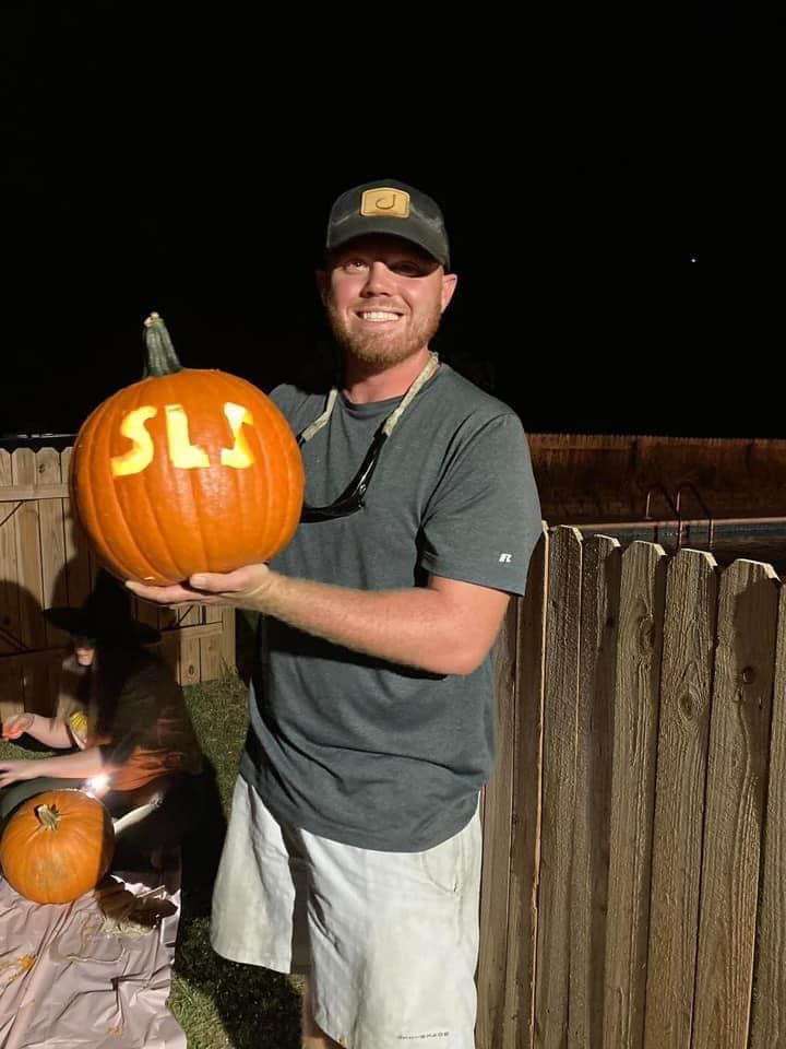 A man is holding a pumpkin with the letters sls carved into it.