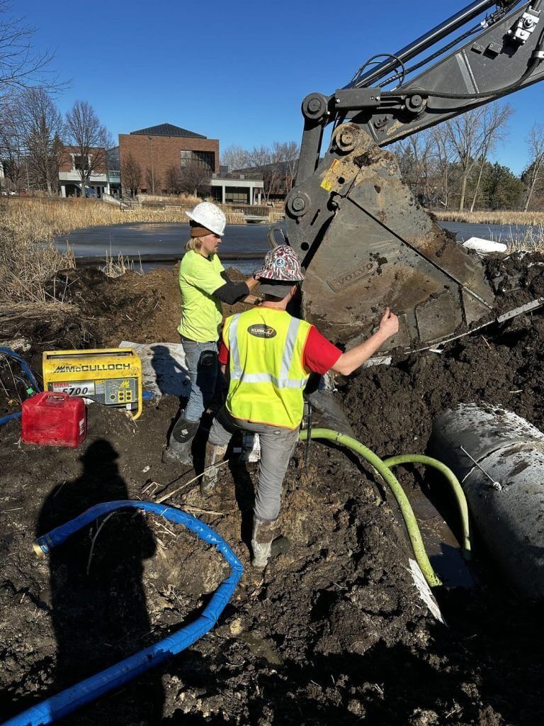 Workers in neon safety vests and hard hats repair a large pipe trench beside an excavator.