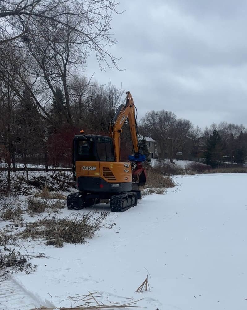 Yellow excavator clearing snow beside a frozen river under a cloudy winter sky