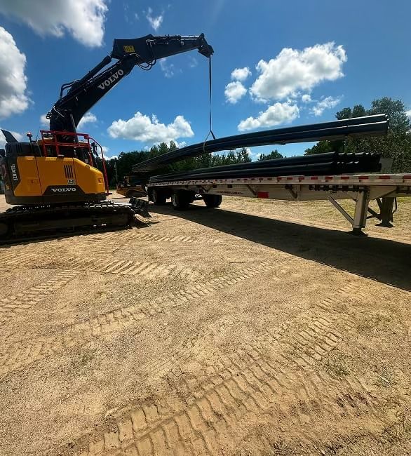 Yellow excavator loading long steel beams onto a flatbed trailer in a dirt yard under a blue sky