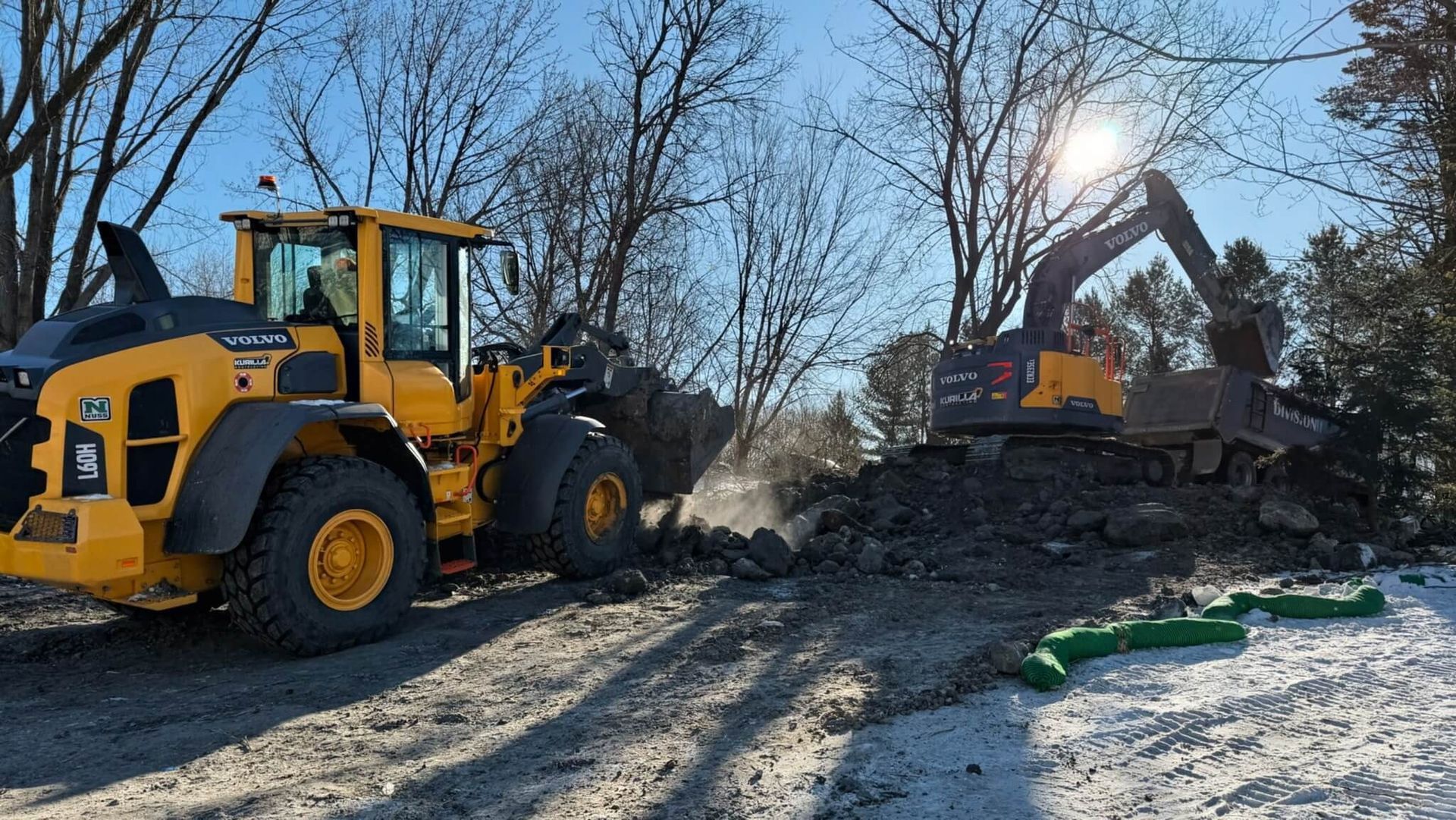 Yellow construction loader and excavator working on a snowy dirt road in a wooded area.