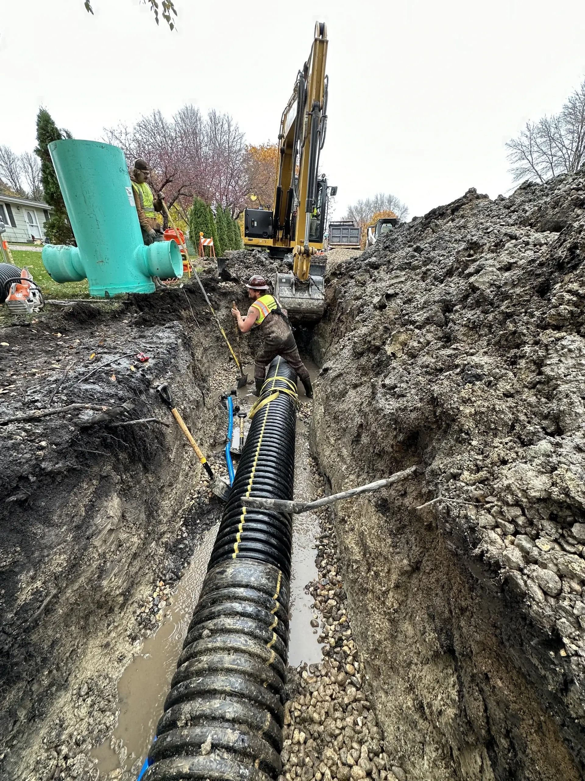 Construction workers laying a black corrugated pipe in a muddy trench beside heavy machinery