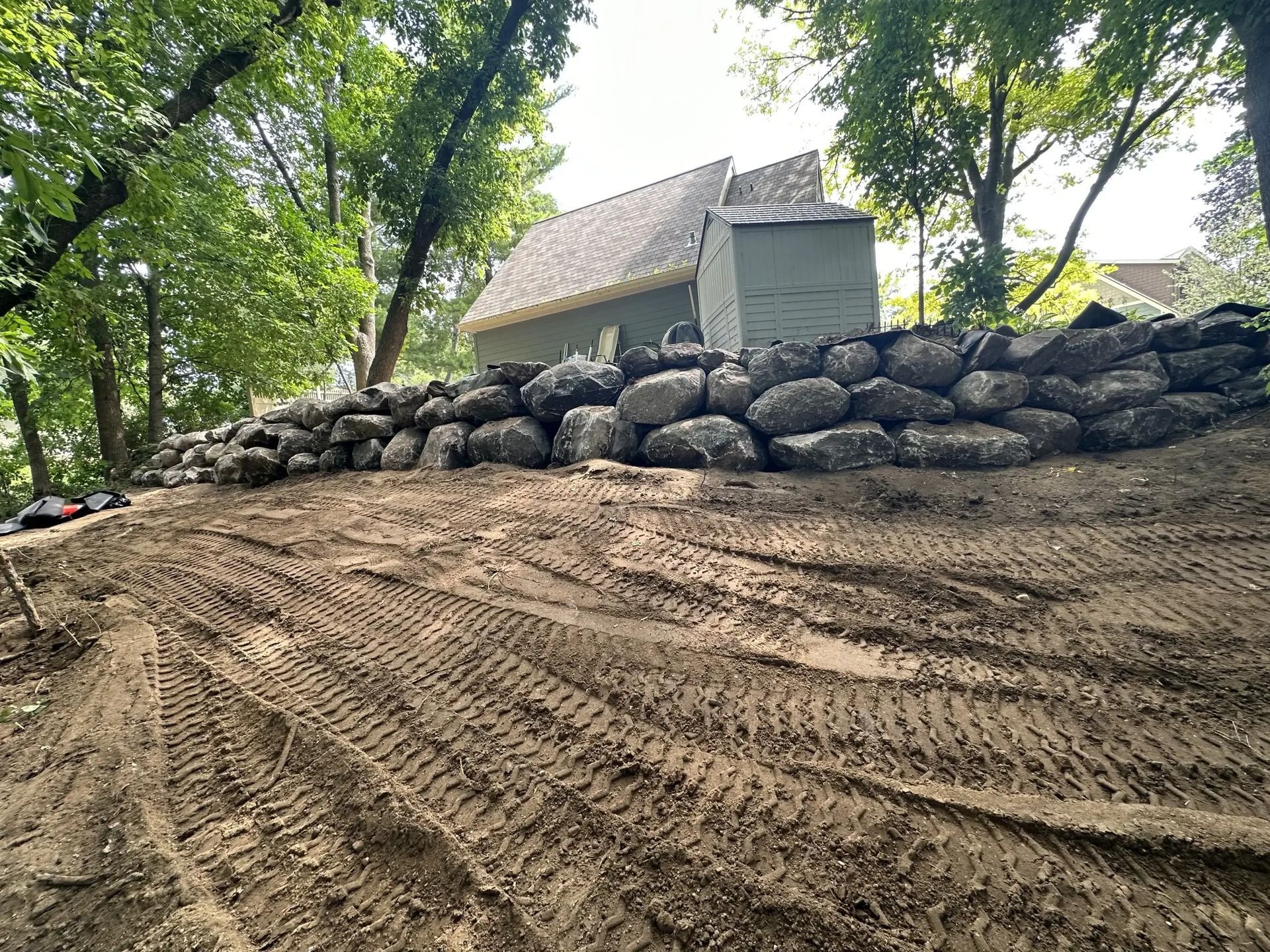 Dirt path beside stacked rocks and trees near a house, with fresh tire tracks in the foreground