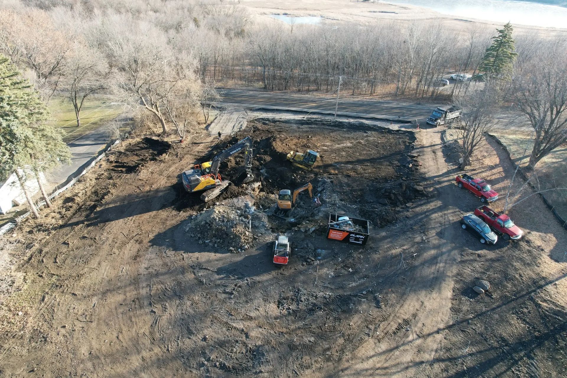 Aerial view of an excavator and several trucks working on a dirt construction site near trees and a road
