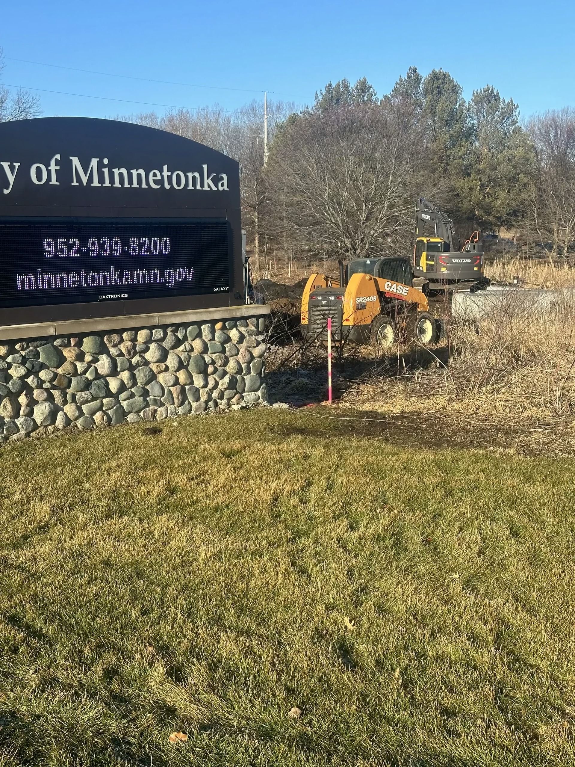 City of Minnetonka sign beside a yellow excavator clearing brush in a grassy area