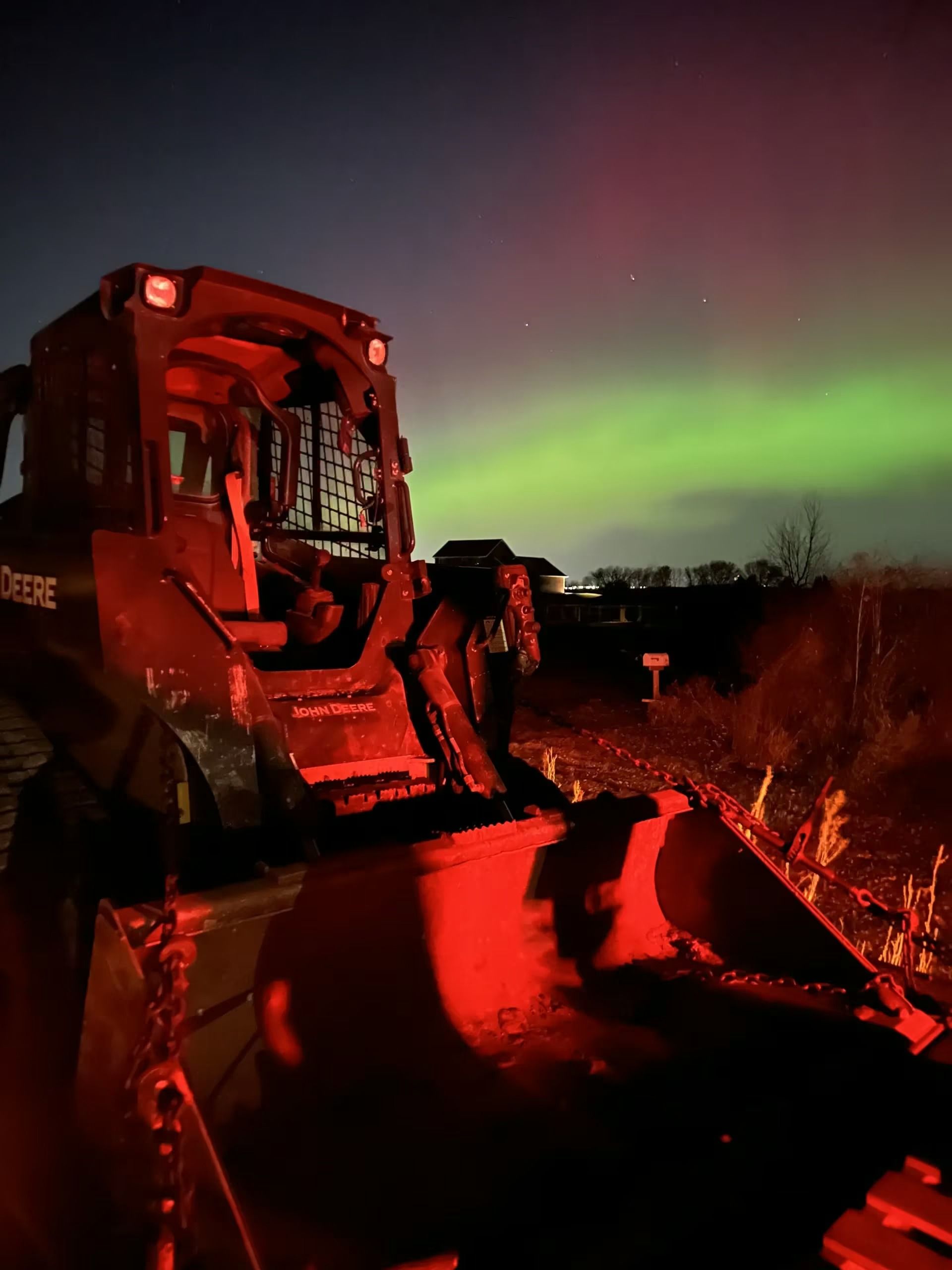 Red snowplow at night under green aurora, with a bright red light illuminating the snow-covered ground.
