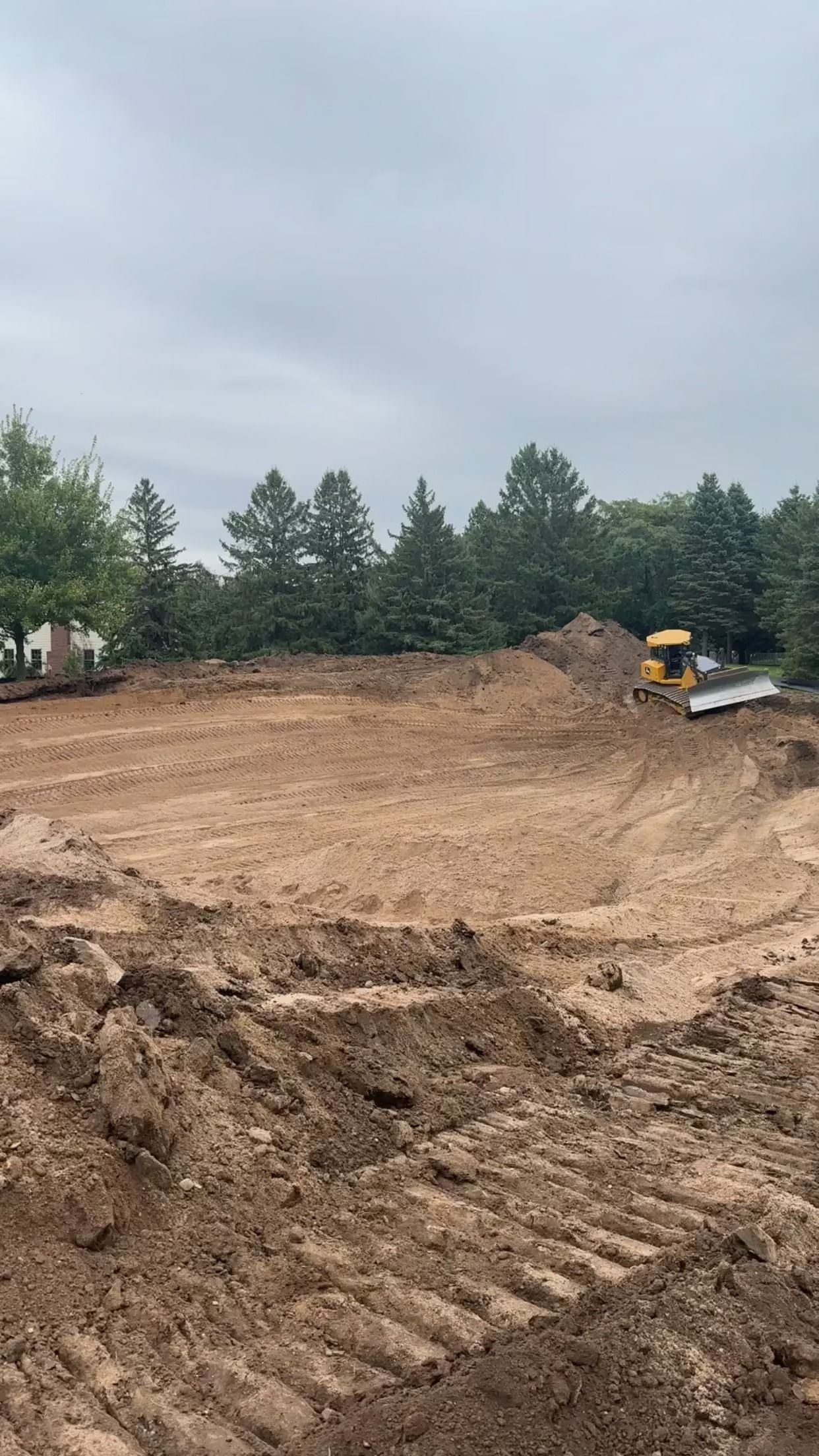 Large muddy construction site with excavated earth, trees in the background, and a small excavator at right.