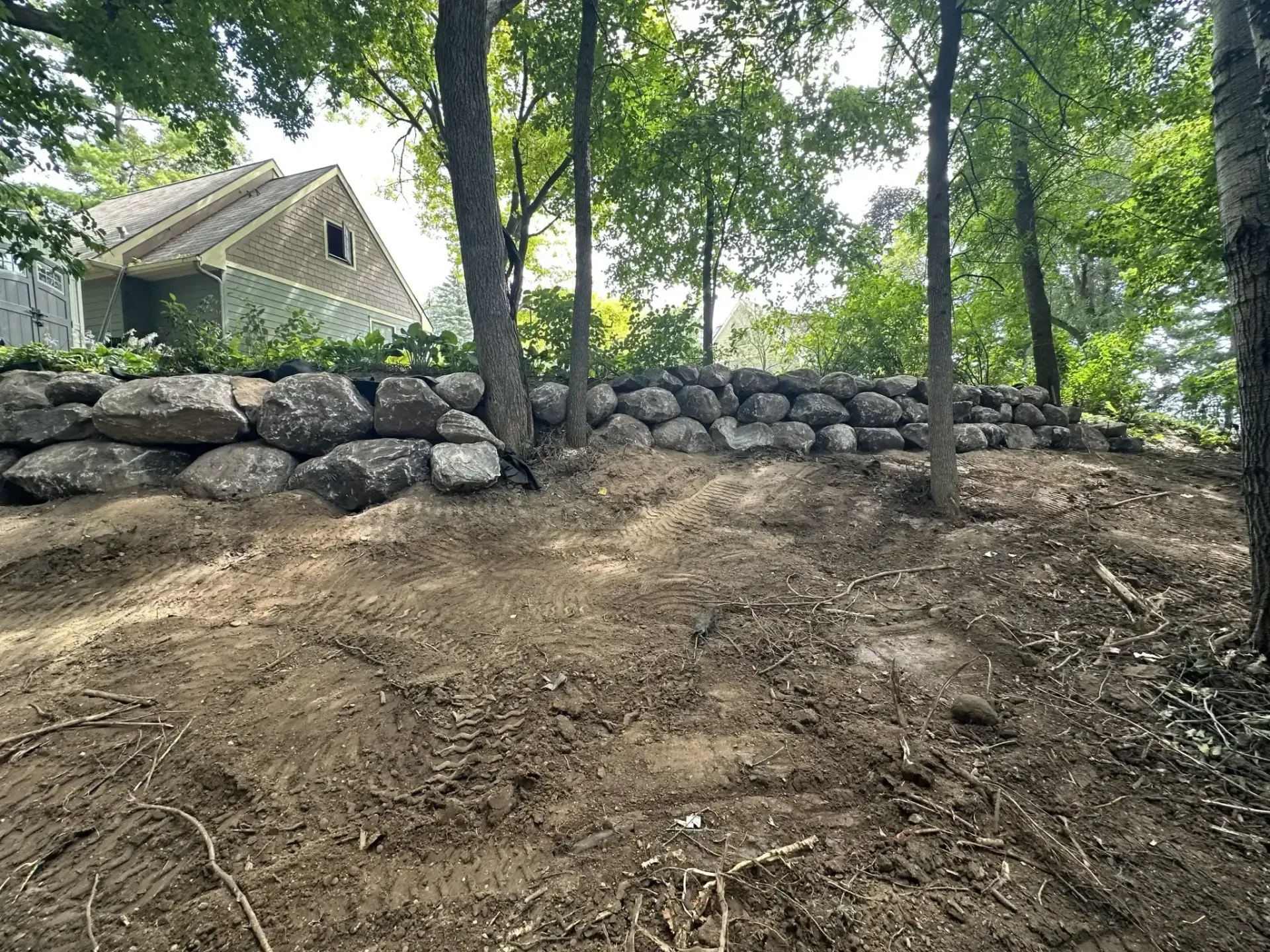 Wooded hillside with a stone retaining wall and a house in the background