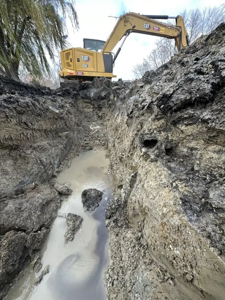 Excavator digging a muddy trench beside a water-filled ditch outdoors