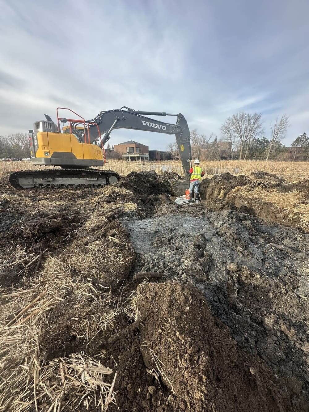 Excavator digging in a muddy field while a worker in a safety vest stands nearby under a cloudy sky