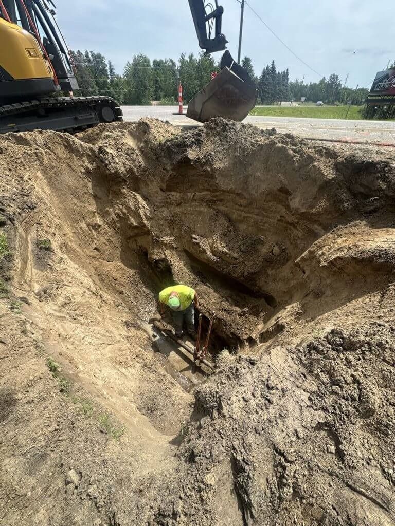 Construction worker in a deep trench beside an excavator bucket at a roadwork site
