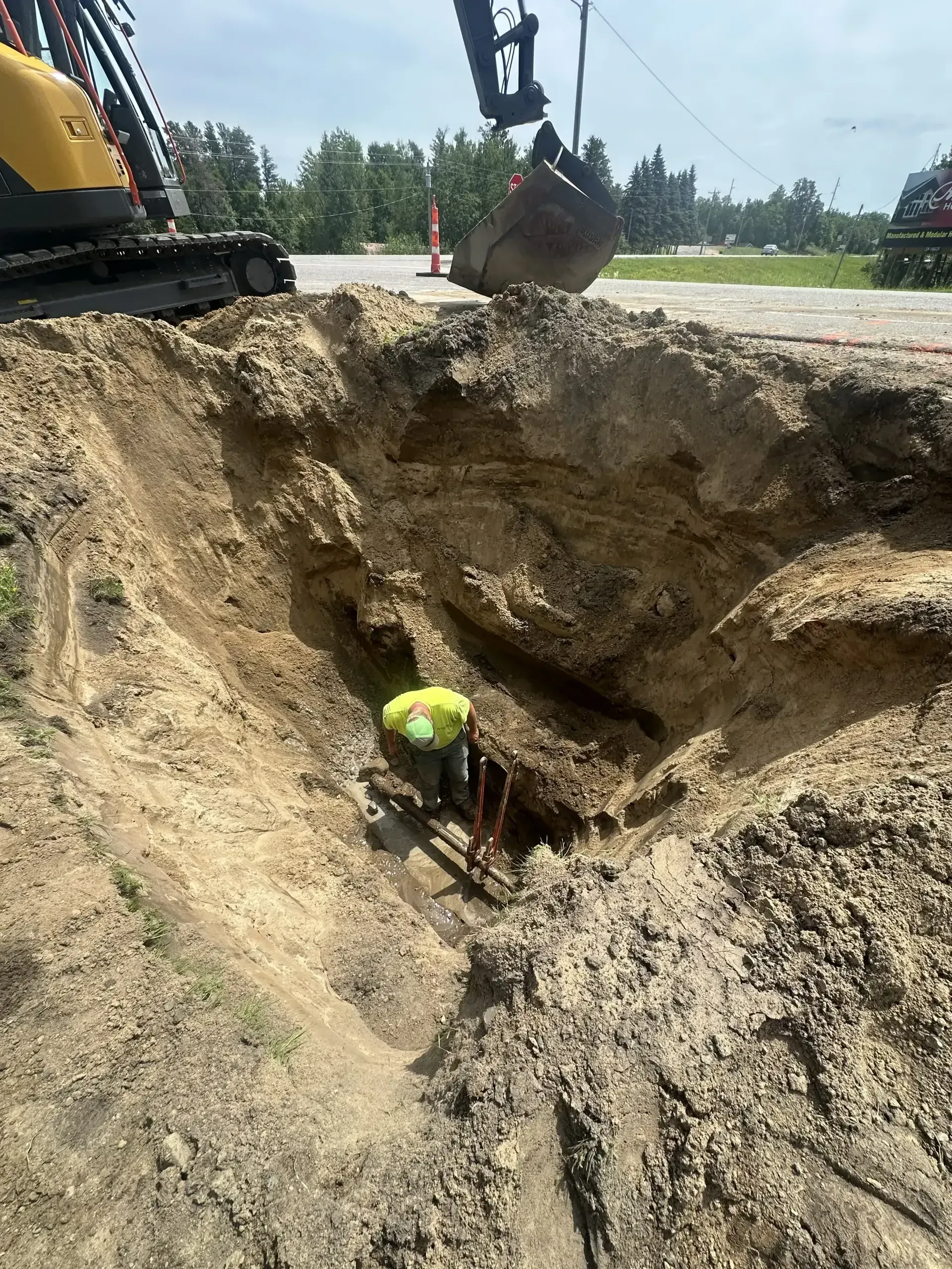 Worker in a yellow hard hat in a deep trench beside an excavator at a construction site.