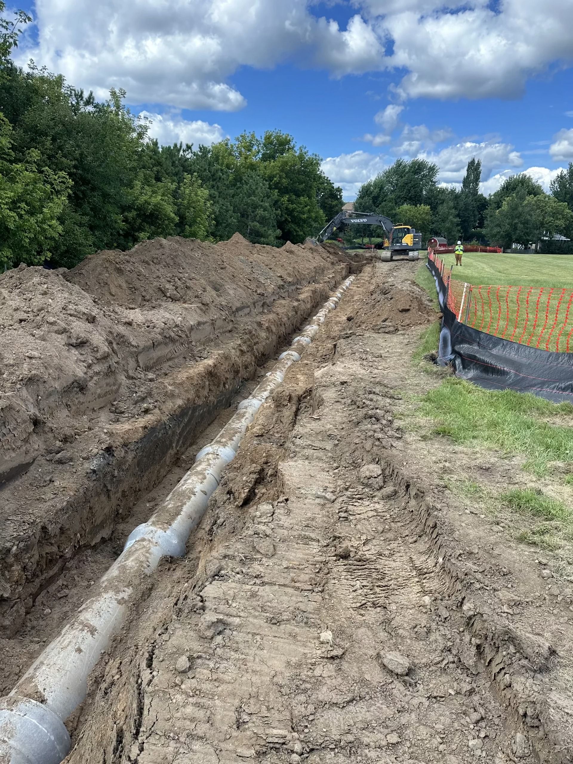Trench with buried pipe alongside a grassy field under a cloudy sky