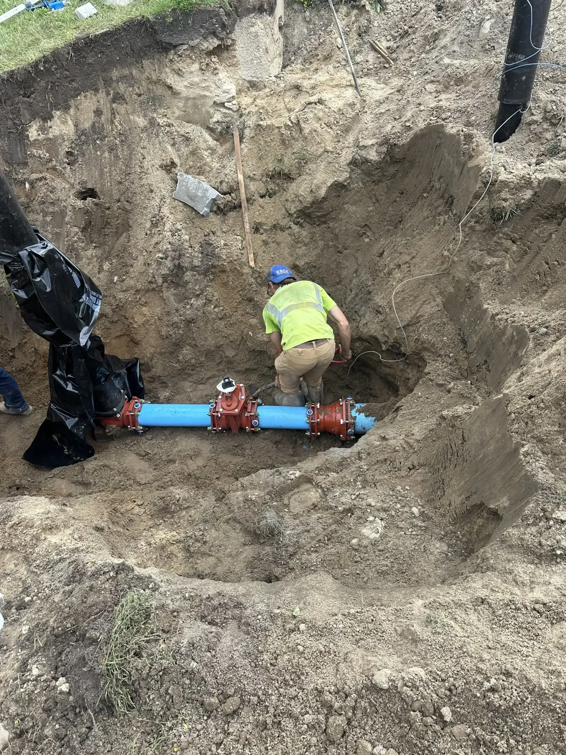 Worker in a neon vest repairs a blue pipe in a deep dirt trench.