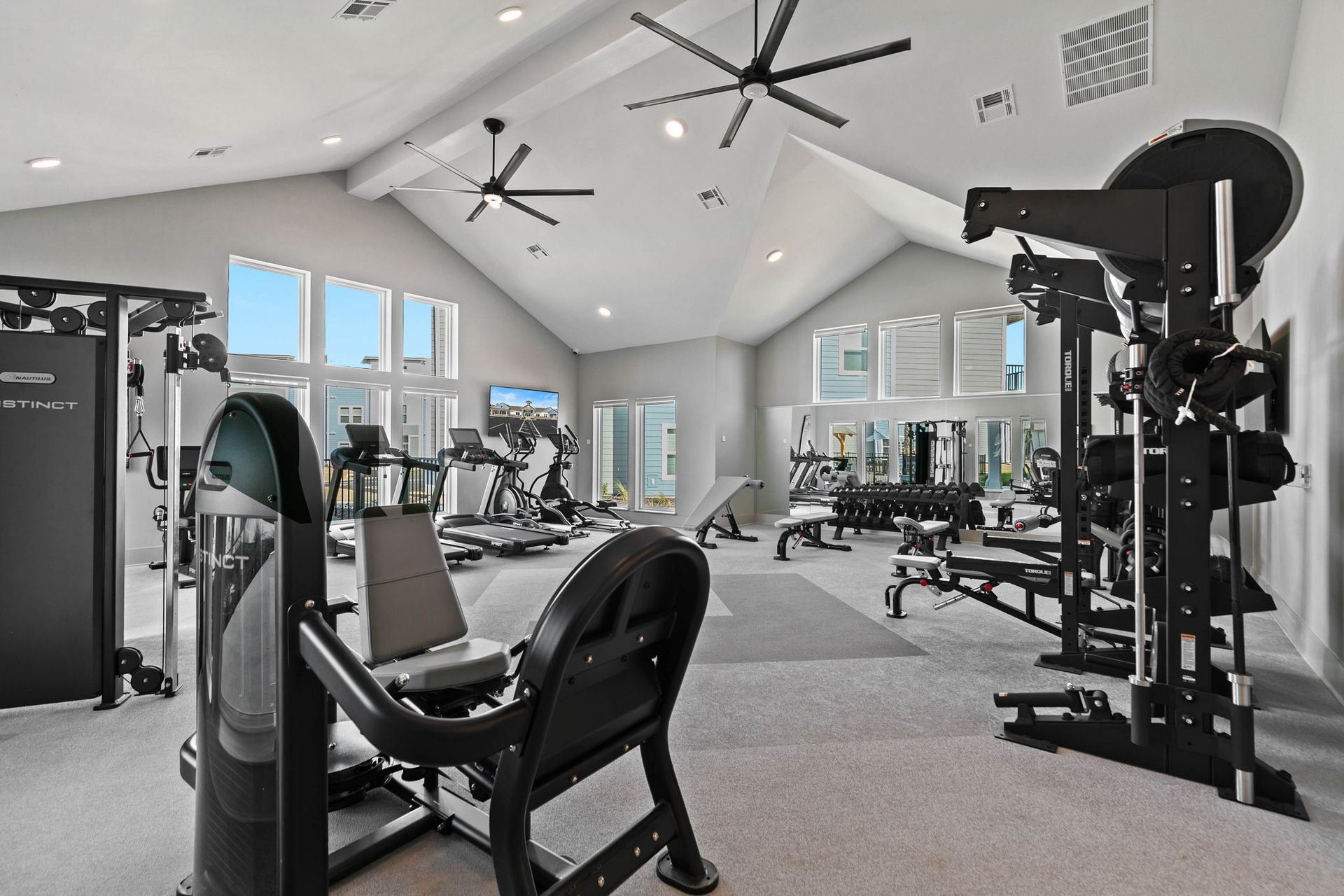 Gym interior with various exercise machines, high ceiling, and large windows.