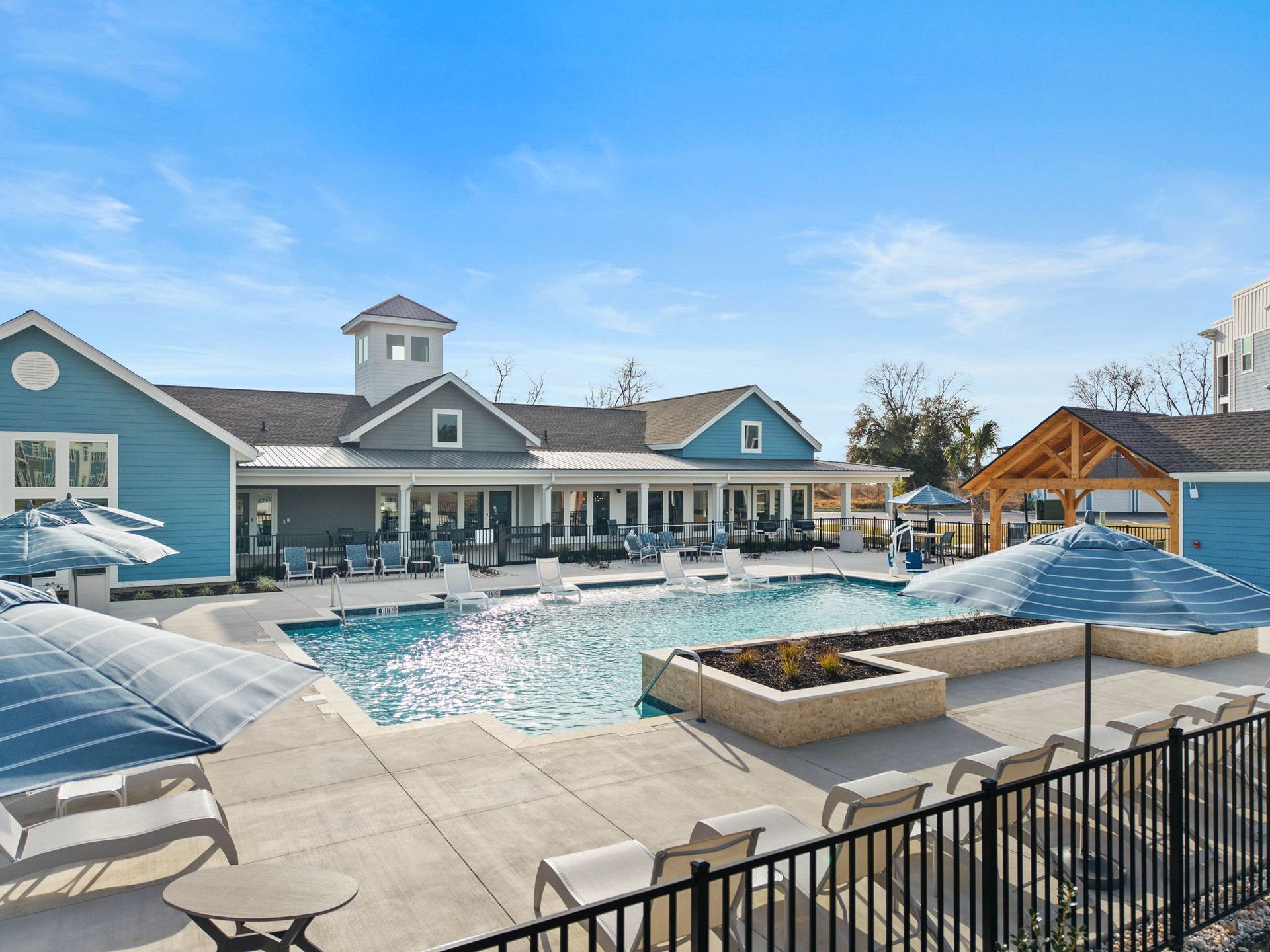 Swimming pool and clubhouse with blue exterior under a clear sky. Sun umbrellas and lounge chairs surround the pool.