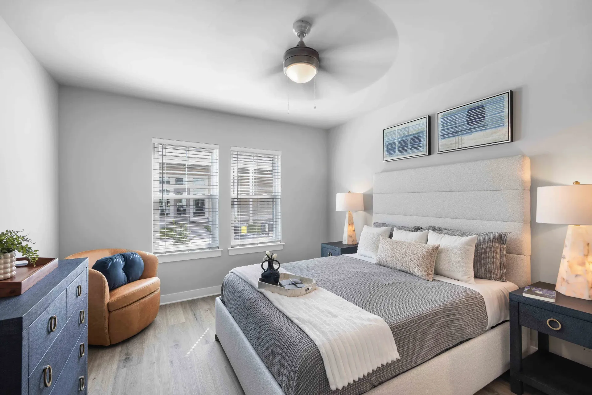 Bedroom with bed, blue chest, and accent chair. Two windows, art, lamps, and a ceiling fan are visible.