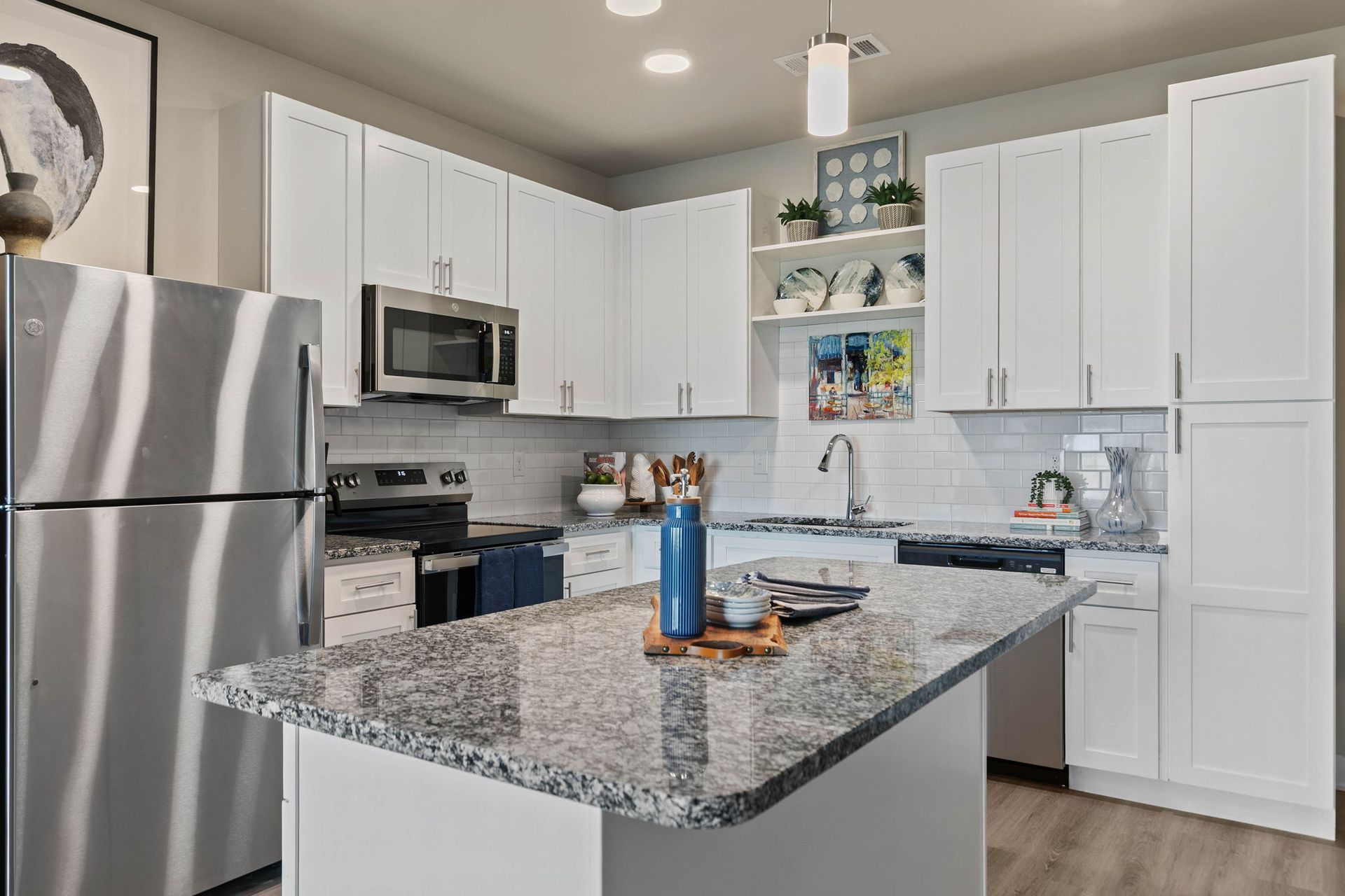 Bedroom with bed, blue chest, and accent chair. Two windows, art, lamps, and a ceiling fan are visible.