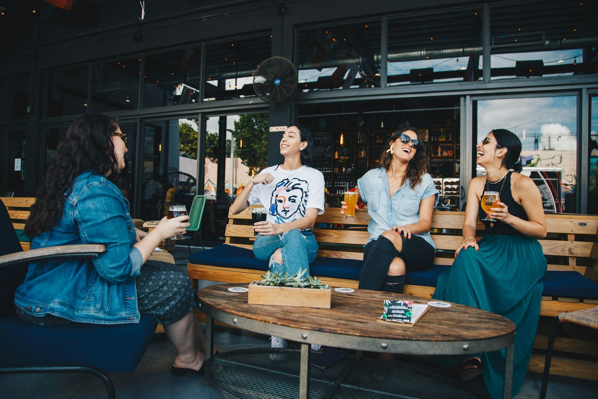 Four people at an outdoor cafe, laughing and holding drinks. Wooden furniture and greenery in a modern setting.